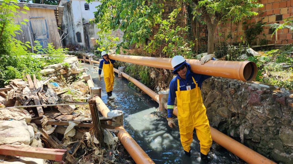 Dois trabalhadores em uniforme amarelo carregam um cano de esgoto grande em uma área com canalização exposta.