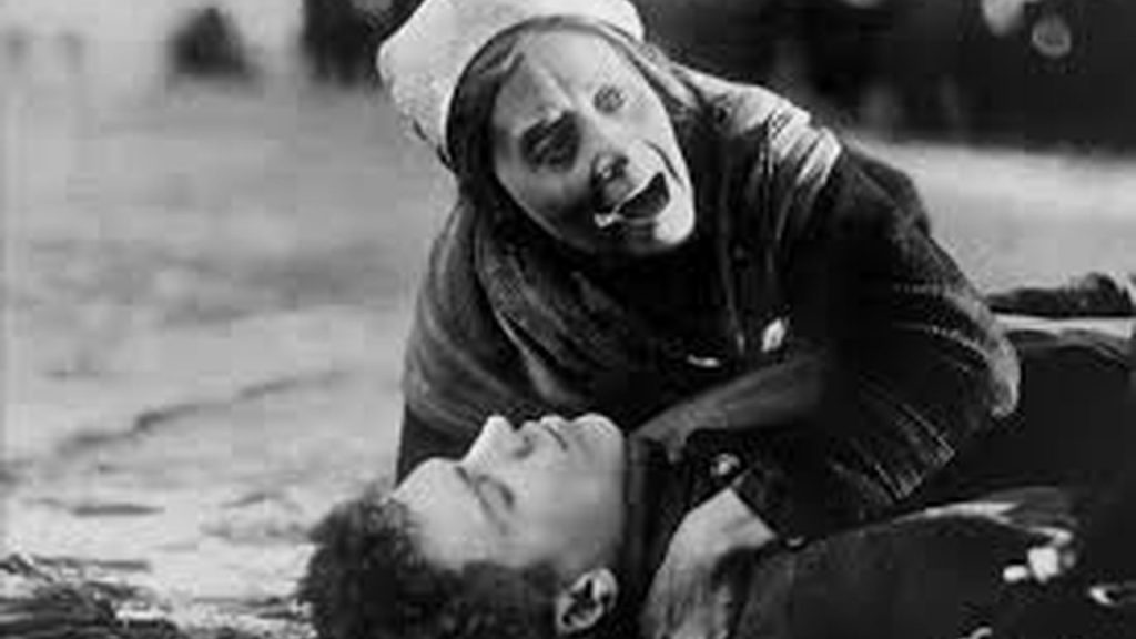 Cena dramática do filme 'A Mãe' (1926), com mulher desesperada e homem ferido.