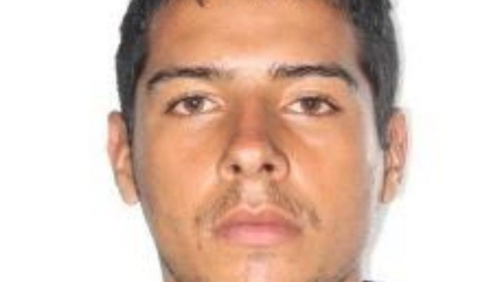 Jovem com pele morena e barba por fazer, em fundo branco.